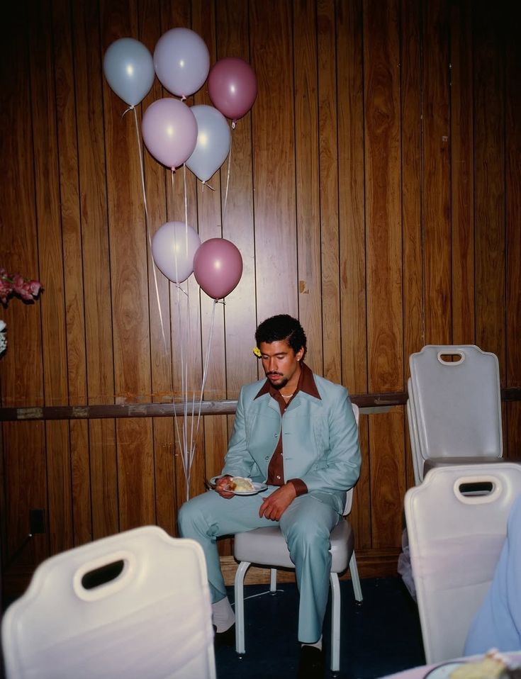 Bad Bunny i blå dress og brun skjorte som sitter på hjørnet og spiser kake med ballonger i bakgrunnen.