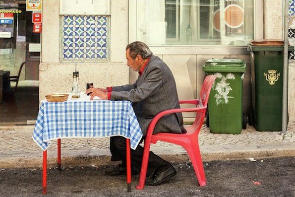 Gammel mann som sitter på en rød monobloc-stol og et matchende bord i plast ute i sidegaten på en restaurant.