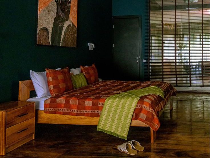 Dobbeltseng med fargerik sengesett, tregulv og stort maleri på veggen av en afrikaner. 